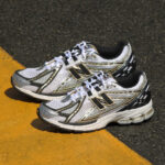 Pair of white and silver running shoes with black detailing resting on a dark asphalt surface next to a yellow line, photographed from above.