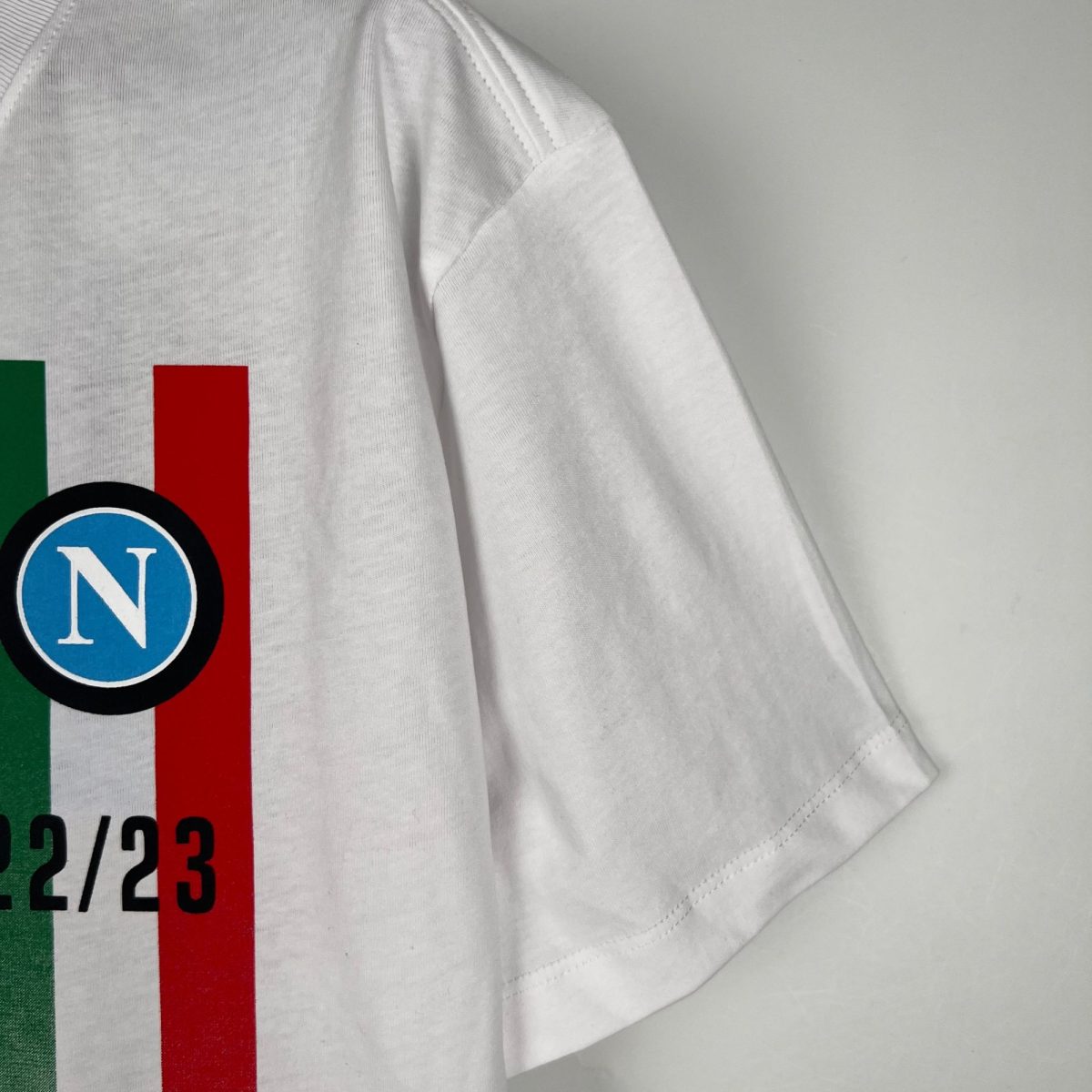 Close-up of a white T-shirt with green and red vertical stripes and a blue circular logo containing a white 'N'. the image shows fabric texture and partial graphic elements near the left side of the shirt.