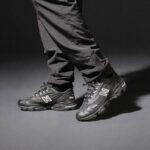 Two black New Balance 990 sneakers on a person wearing dark jeans in a studio setting with a grey background.