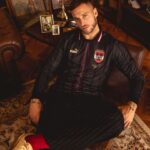 Man seated in a living room wearing a black pinstripe soccer jersey with a crest, indoors by a coffee table with framed photos behind him.