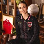 Portrait of a smiling woman wearing a black pinstripe soccer jersey with a crest, indoors among books and memorabilia including a globe and framed red jersey in the background, conveying a casual, sporty vibe.