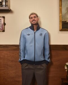 Man wearing a light blue Adidas track jacket with dark cuffs and stripes, standing against a wood-paneled wall indoors.