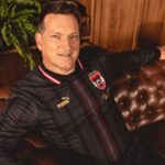 Man in a black pinstripe Puma football jersey sitting on a brown leather sofa with a wooden wall and fern in the background.