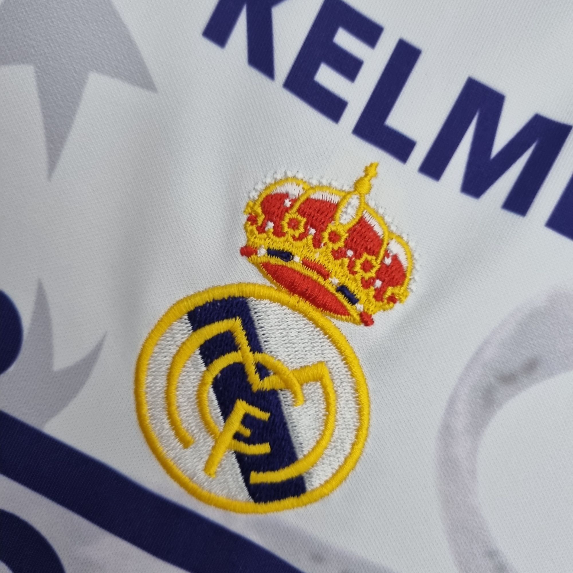 467e8761_1789cfe7-a44e-4306-a8c8-bf67f1635c80 | UAE SPORT STORE Close-up of an embroidered emblem on white fabric, showing a crown-topped shield in red and yellow with blue detailing and large blue lettering nearby.