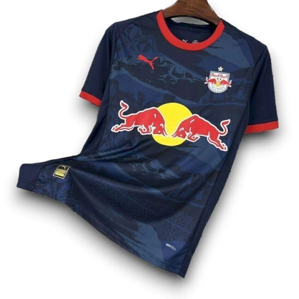 Navy blue soccer jersey with red trim on the sleeves, hanging on a wooden hanger; features a large Red Bull logo (two red bulls charging toward a yellow circle) across the chest and a small team badge on the left chest.