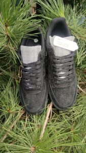 Pair of black sneakers with gray tongues resting on pine needles, a tag attached to the left shoe visible in the scene.
