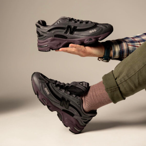 Person holding a gray and purple New Balance 1000 sneaker mid-air, beige background visible.