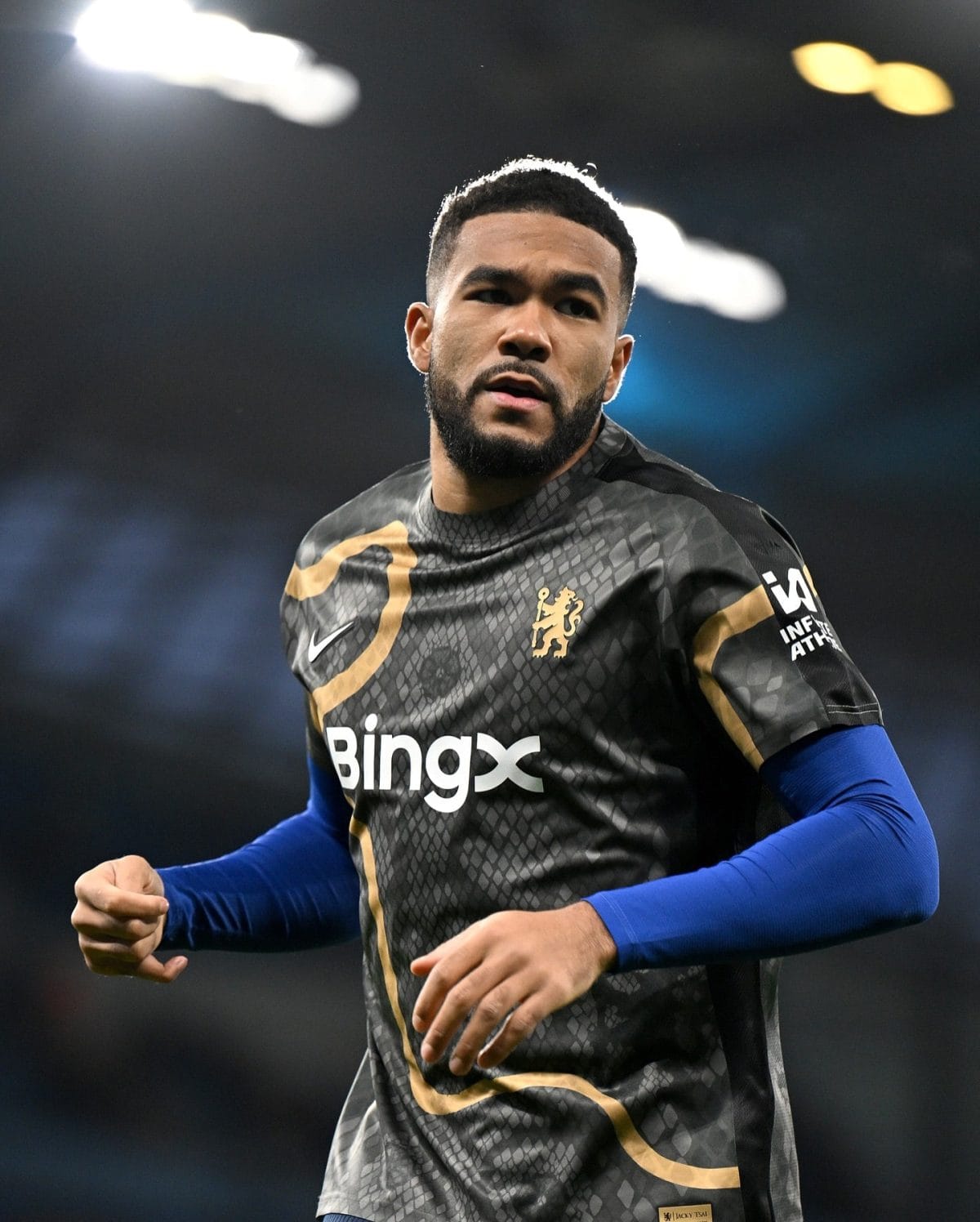 Soccer player in a dark patterned jersey running on the field, blue undershirt visible at the arms, focused expression.