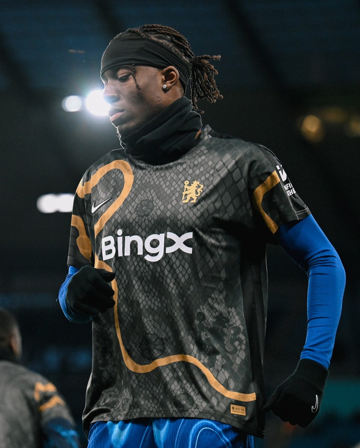 Soccer player in a black patterned jersey and blue undershirt, standing on the field with a headband and ruffled hair under stadium lights, looking downward.