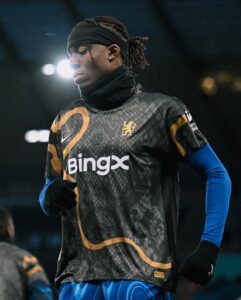 474937609_18492982984021107_5853922922866389082_n | UAE SPORT STORE Soccer player in a black patterned jersey and blue undershirt, standing on the field with a headband and ruffled hair under stadium lights, looking downward.