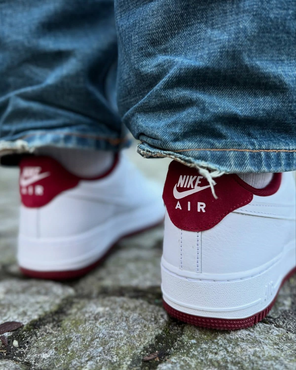 Close-up of white Nike Air sneakers with red heel tabs peeking from denim jeans outdoors on a rough stone surface