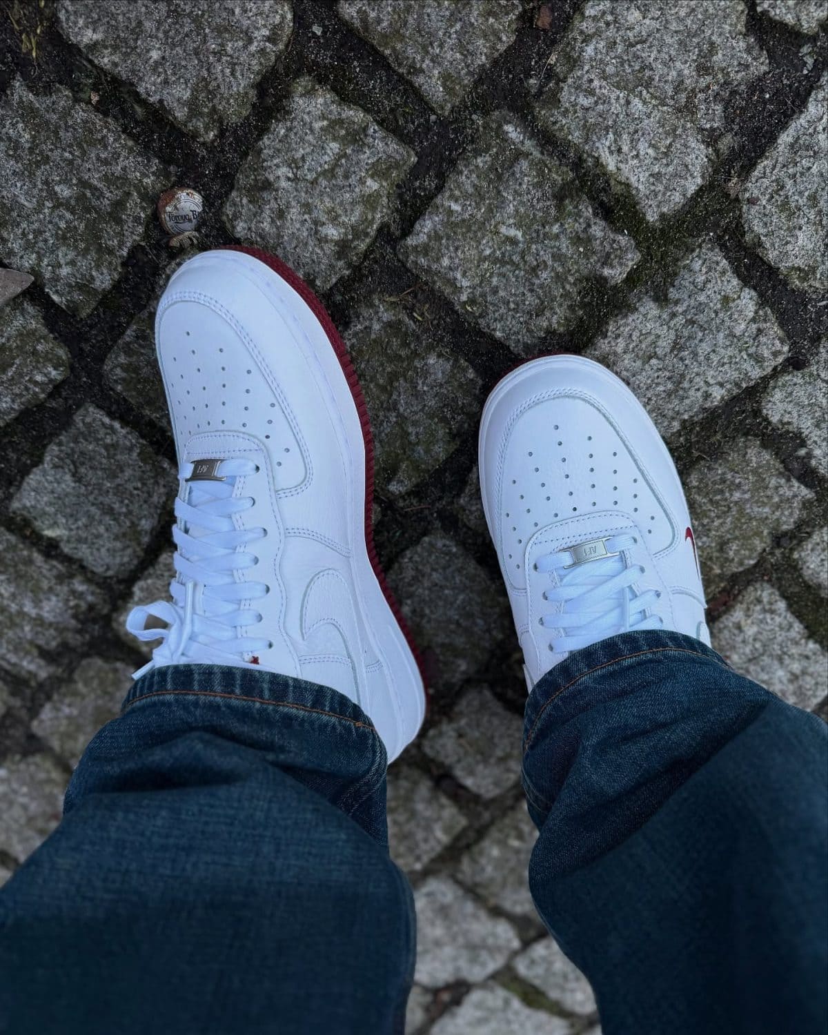 Person wearing white sneakers with red soles standing on rough cobblestone ground, jeans cuffs visible.