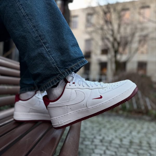 Close-up of white Nike sneakers with red swoosh resting on a brown bench, denim jeans visible at the ankle, outdoor urban setting behind them.