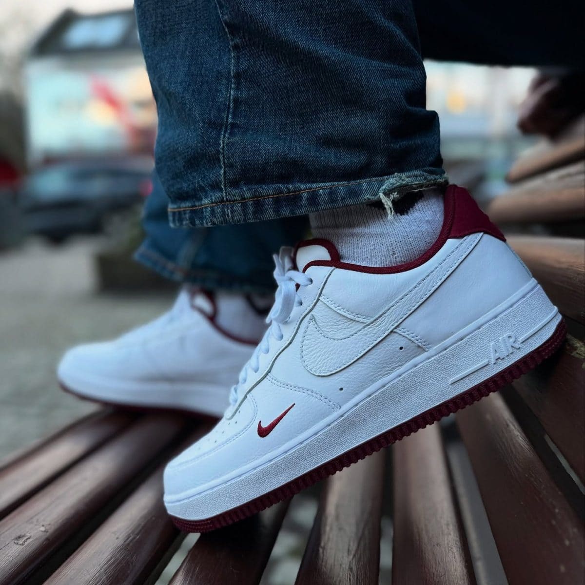 Close-up of white Nike Air sneakers with burgundy accents resting on a wooden bench, denim jeans cuffed above.