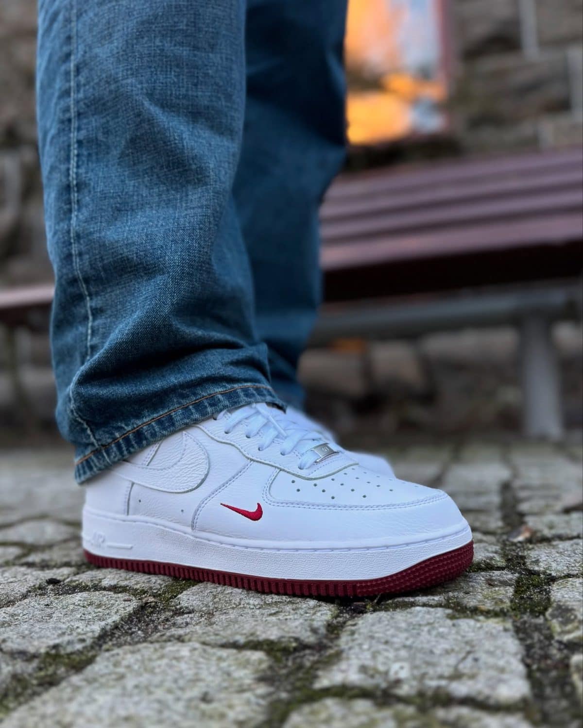 White Nike Air Force 1 sneaker with a red swoosh on a stone pavement, paired with blue jeans and a visible denim hem.
