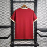 Red short-sleeve sports jersey hanging on a wooden hanger against a metal rack, back view with cream collar and cuffs.