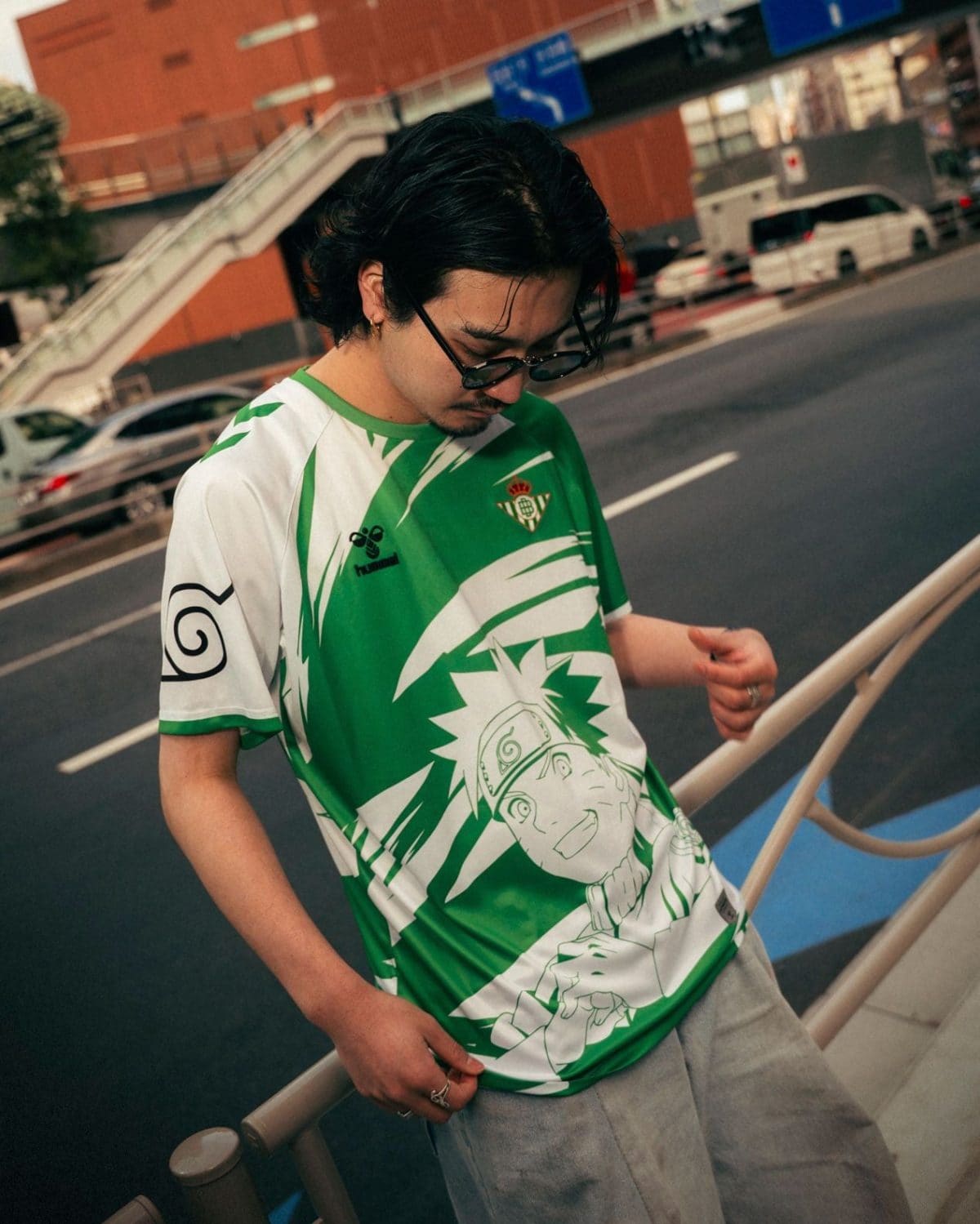 Young man in a green Naruto graphic tee leaning on a railing outdoors, looking down at his hand with sunglasses on.