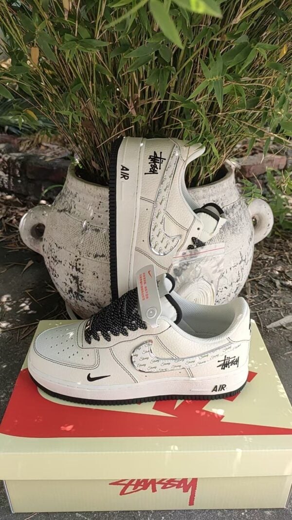 485e9a5a | UAE SPORT STORE White Nike Air Force 1 sneakers with Stussy branding resting on a pale shoebox beside a potted plant.