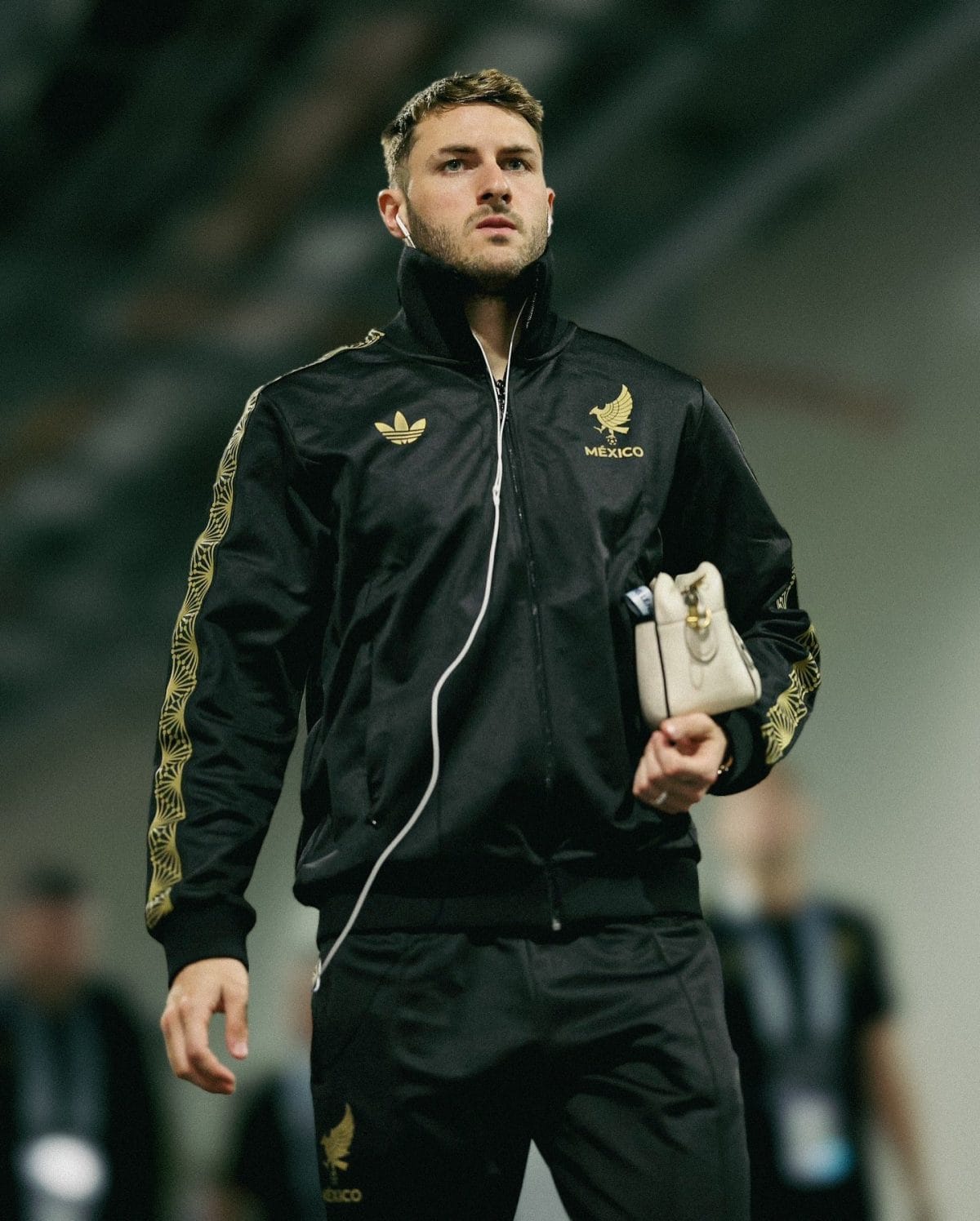 Athlete in a black Mexico jacket with gold emblem, wearing white earbuds, holding a small beige bag.