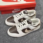 Pair of white and brown Nike sneakers with a brown Nike swoosh, placed next to a red Nike shoebox on a grey carpet.