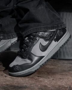 Close-up of a black and gray Nike sneaker with a visible swoosh, worn with dark jeans, on a concrete floor.