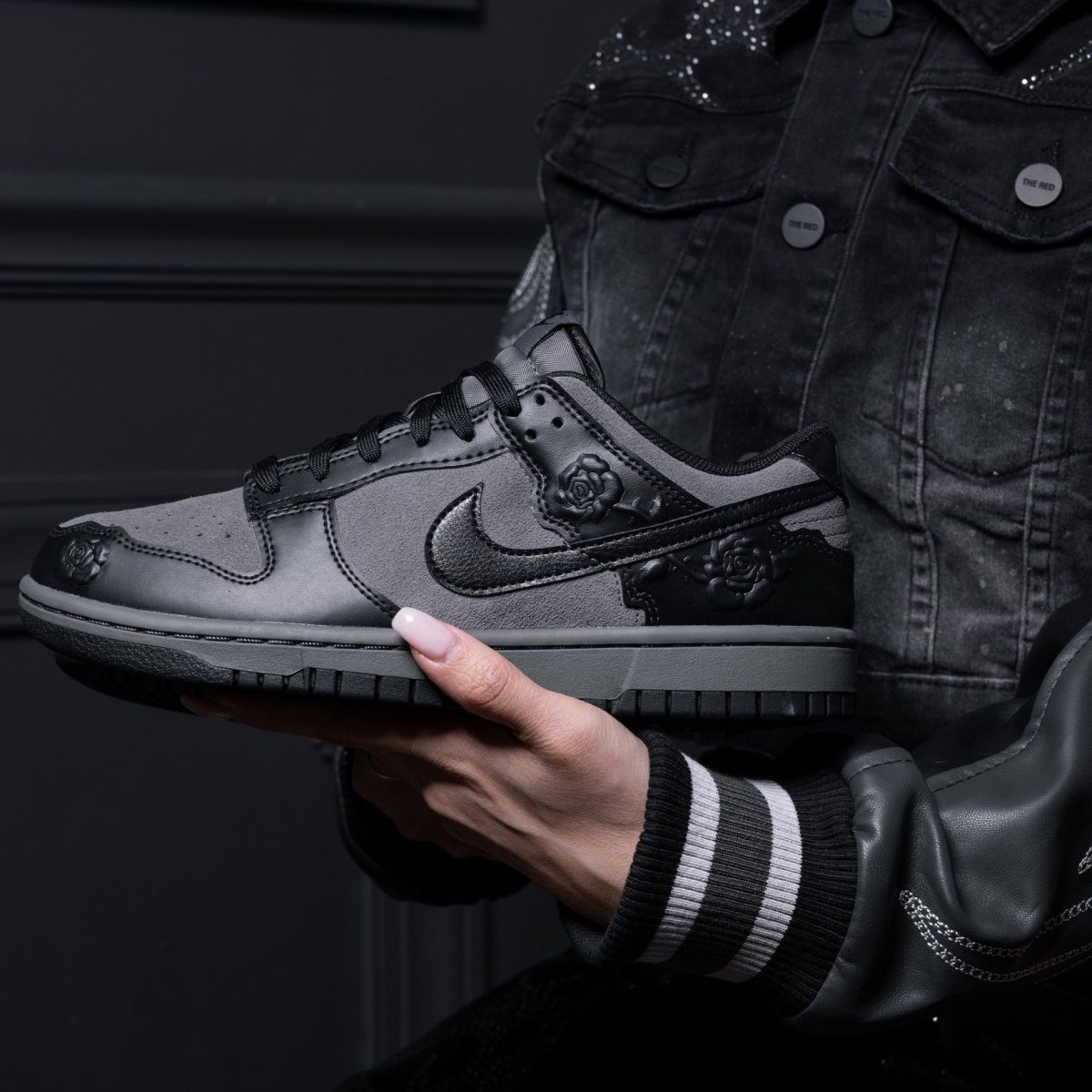 Person in a dark denim jacket holds a gray-and-black sneaker with rose embellishments against a dark background.