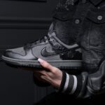 Person in a dark denim jacket holds a gray-and-black sneaker with rose embellishments against a dark background.
