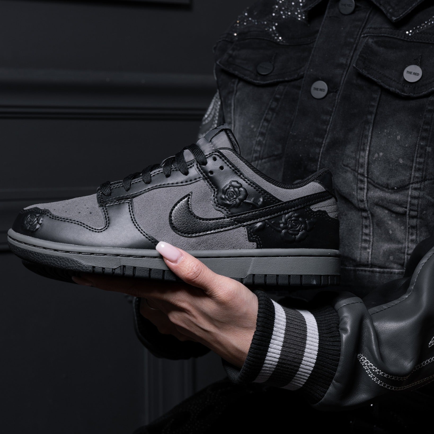 490106728_1104037408425707_7546062657029446279_n Person in a dark denim jacket holds a gray-and-black sneaker with rose embellishments against a dark background.