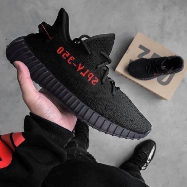 Close-up of a black knit sneaker with red lettering along the side, being held over a gray floor; a shoebox is in the background.