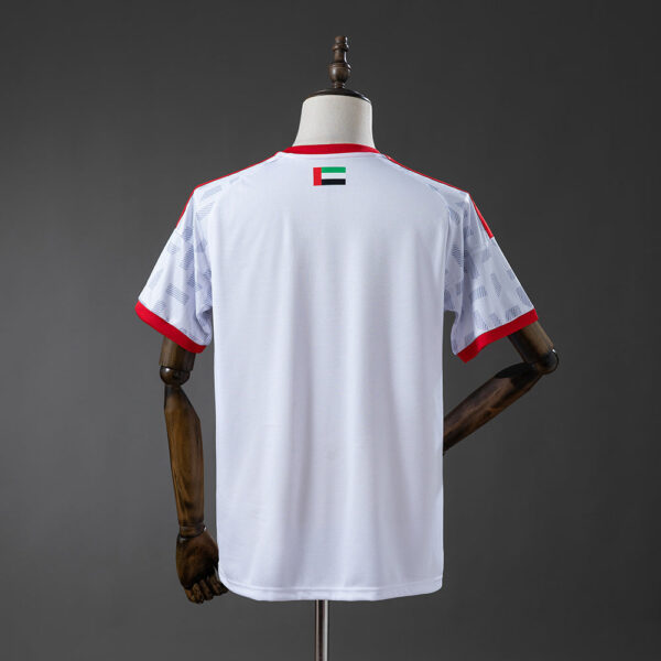 White athletic jersey on a mannequin, red collar and sleeve cuffs, small UAE flag patch on the upper back.