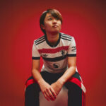 Person seated on a white stool against a vivid red background, wearing a white jersey with black and red stripes and sponsor logos, looking upward to the left.