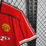 Red Manchester United football jersey hanging on a black metal rack with a diamond-mesh background, crest visible on the chest area.
