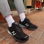 4a0839fa-a568-4361-8ec4-da94a7f97b7f_28d6bcf0-52cf-4fa0-9828-6063fa6eb3fb | UAE SPORT STORE Person wearing black New Balance sneakers with white logo, white socks, and gray plaid trousers standing on a brick sidewalk outside a shop.