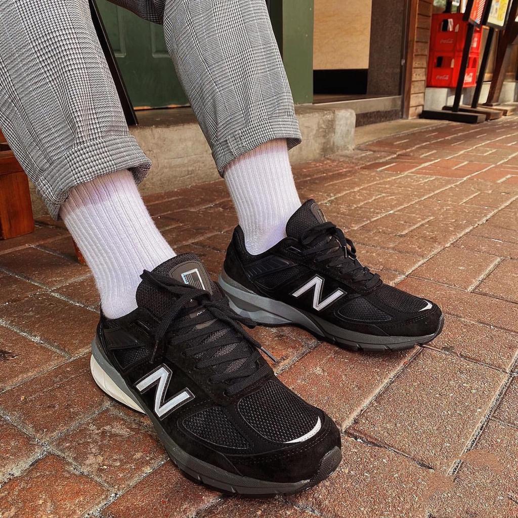 4a0839fa-a568-4361-8ec4-da94a7f97b7f_28d6bcf0-52cf-4fa0-9828-6063fa6eb3fb | UAE SPORT STORE Person wearing black New Balance sneakers with white logo, white socks, and gray plaid trousers standing on a brick sidewalk outside a shop.