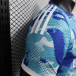 Close-up of a blue camouflage-style athletic jersey with three white Adidas stripes on the shoulder, worn by a person beside a mesh wall.
