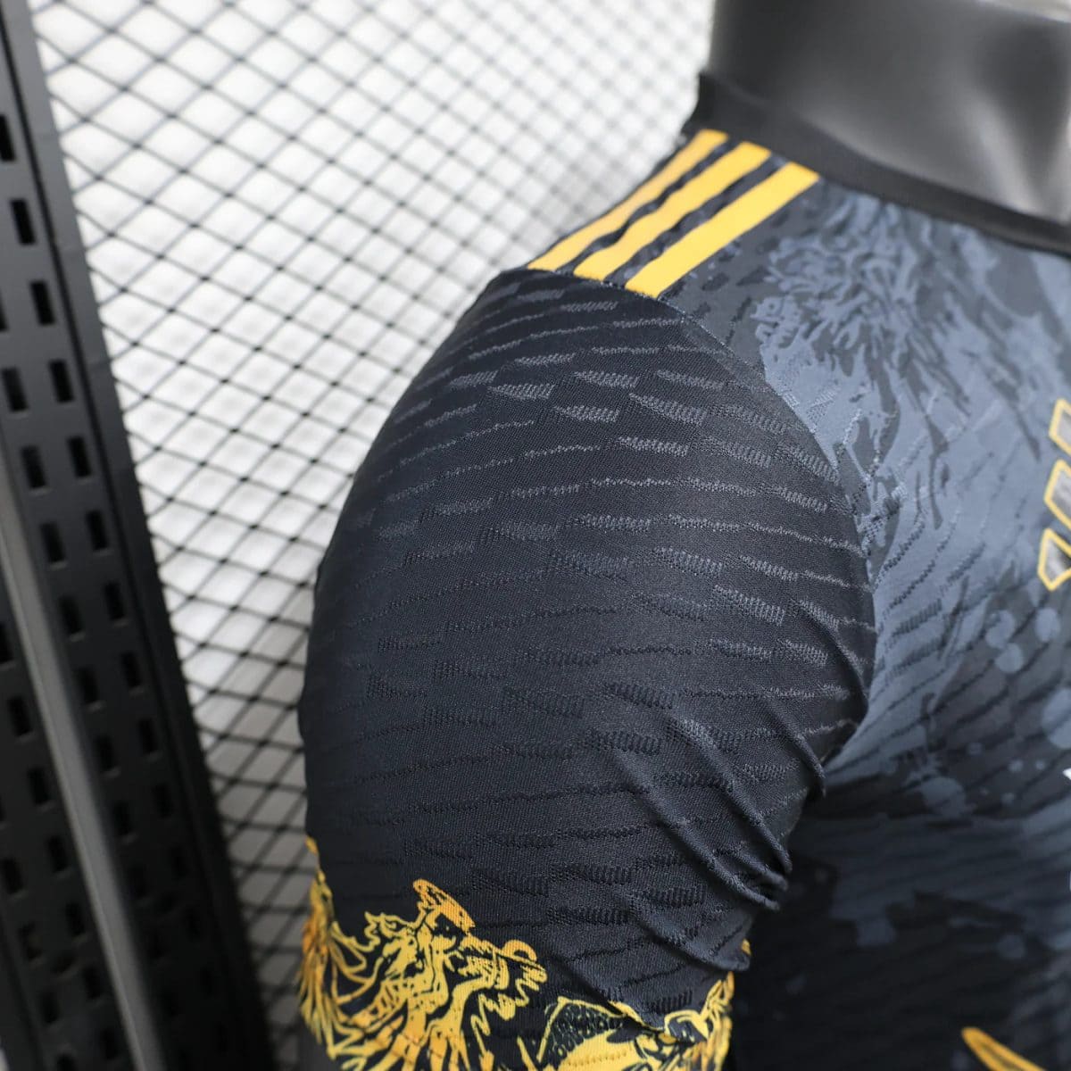 Close-up of a black athletic jersey sleeve with a yellow dragon graphic and three yellow shoulder stripes against a metal grid background.