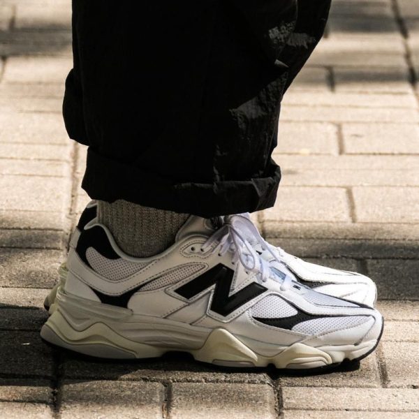 White athletic sneaker with black detailing on a gray sock, worn with rolled black pants on a paved sidewalk.