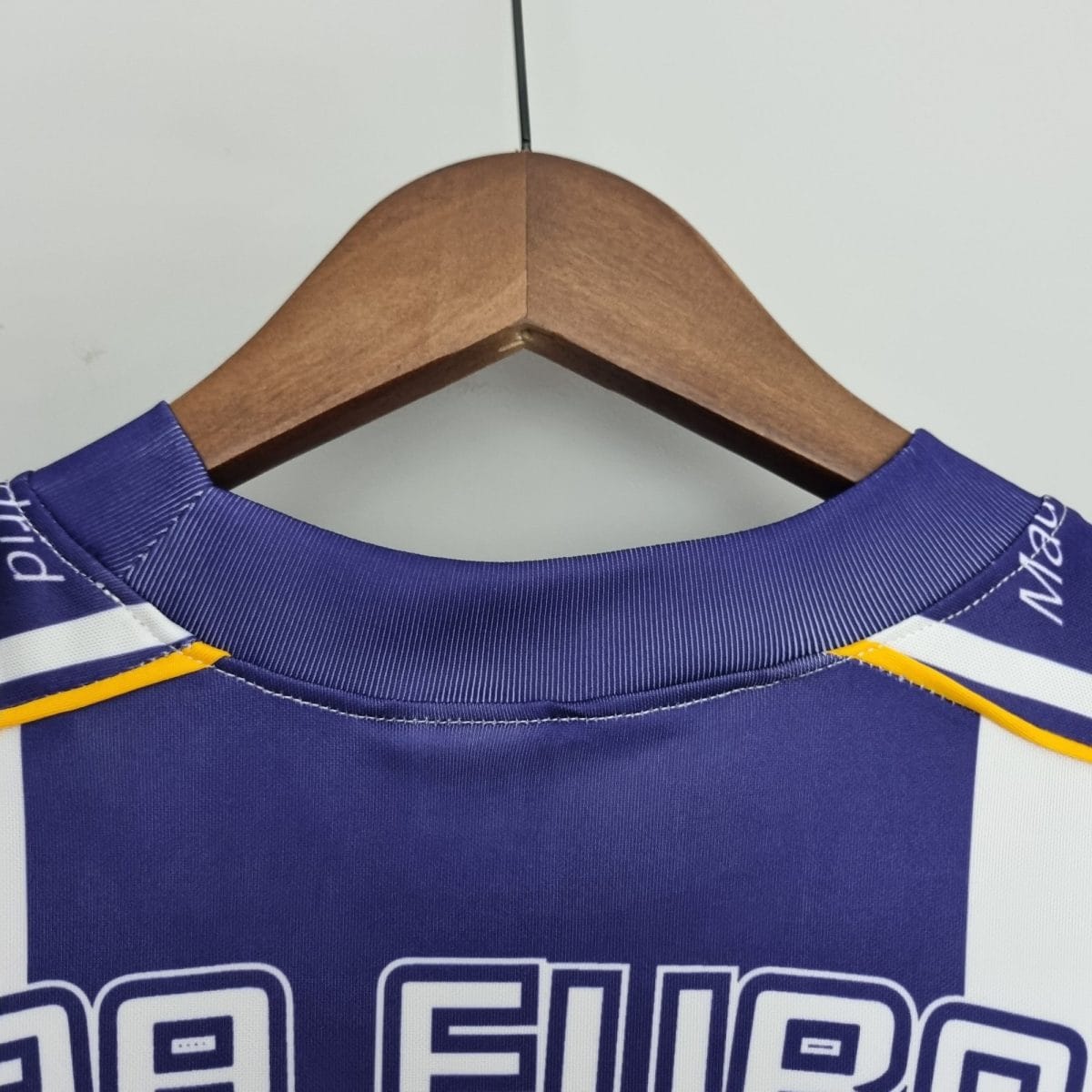 Wooden clothes hanger with a blue polo shirt draped over it, hanging against a plain light background; close-up at the neck showing stitching and a small yellow accent.
