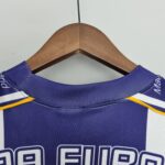 4dcf1889_e41bd504-531a-4c53-a10c-a671e4df72f9 | UAE SPORT STORE Wooden clothes hanger with a blue polo shirt draped over it, hanging against a plain light background; close-up at the neck showing stitching and a small yellow accent.