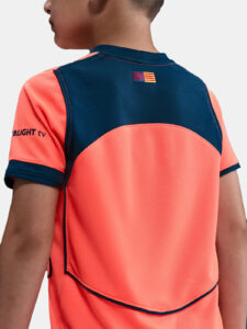 Back view of a child wearing a coral and navy sports jersey with a small striped flag patch on the upper back.
