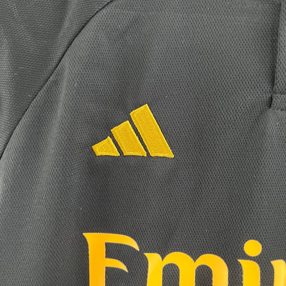 Close-up of dark knit fabric with a yellow embroidered three-bar logo on the left side of the image.