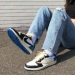 Person sitting on pavement with legs crossed, wearing ripped jeans and white, black, and blue sneakers (Nike).