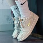 White Nike sneakers with a gold swoosh, paired with light blue socks, resting on a dark floor.