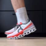 Person wearing a white and orange running shoe with a unique segmented sole, stepping forward on a wooden floor; 'RUNNING' text along the heel.