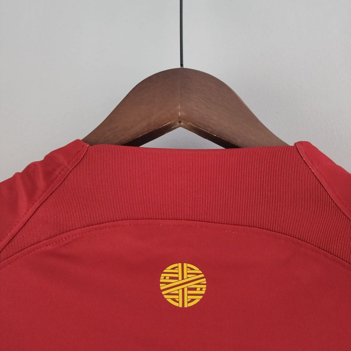 Red garment on a wooden hanger with a metal hook against a light wall; close-up of the upper chest area and stitching visible.