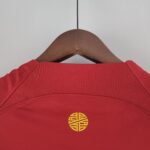 4f9a81f4_fca3bf2f-b9fa-4bca-aaaa-9c8e34d167e7 | UAE SPORT STORE Red garment on a wooden hanger with a metal hook against a light wall; close-up of the upper chest area and stitching visible.