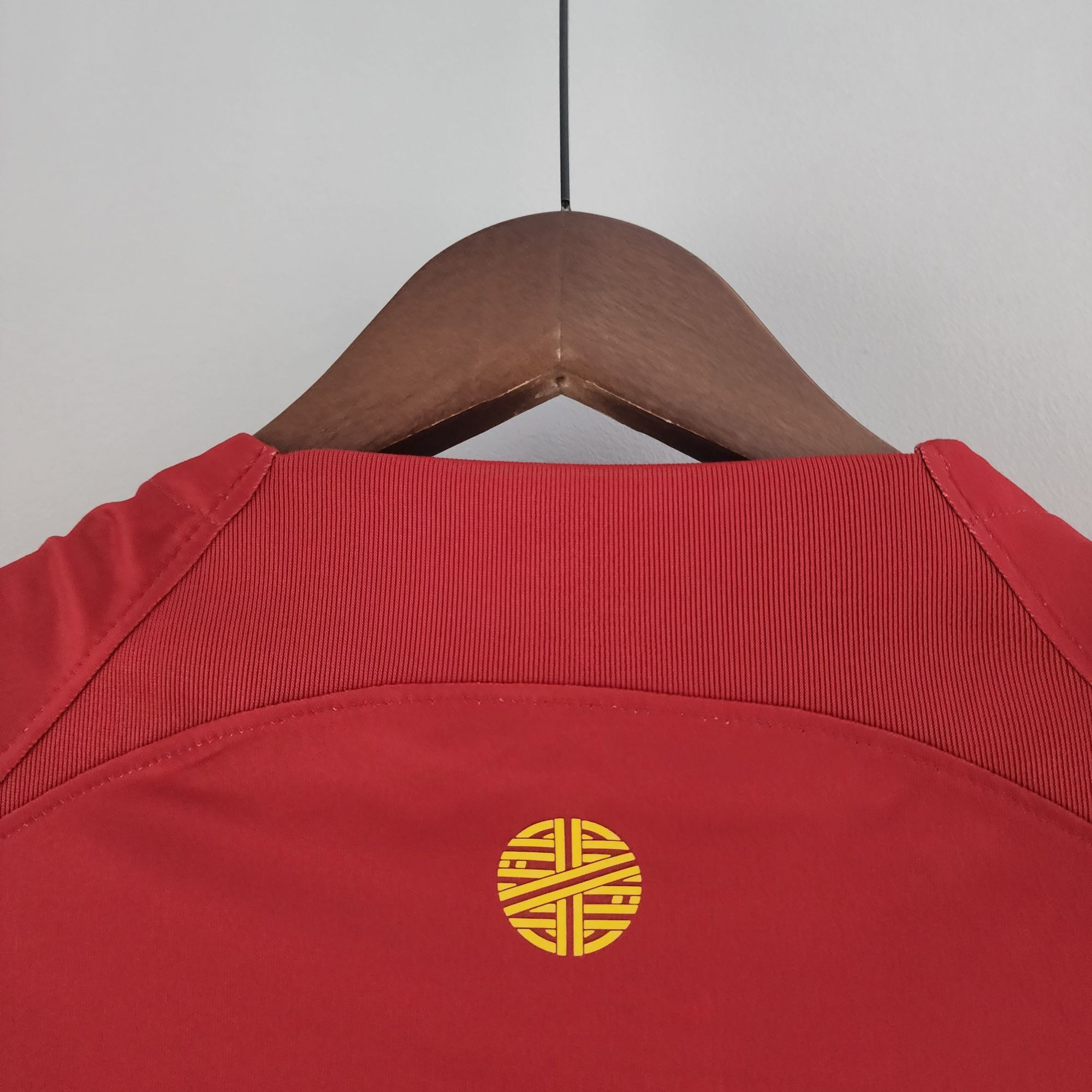 4f9a81f4_fca3bf2f-b9fa-4bca-aaaa-9c8e34d167e7 | UAE SPORT STORE Red garment on a wooden hanger with a metal hook against a light wall; close-up of the upper chest area and stitching visible.
