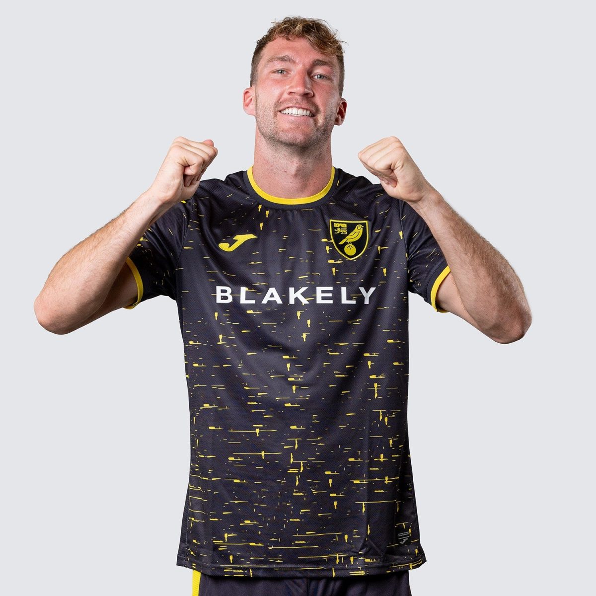 Male soccer player in a navy-yellow kit raises fists in celebration, smiling at camera, team crest visible on chest.