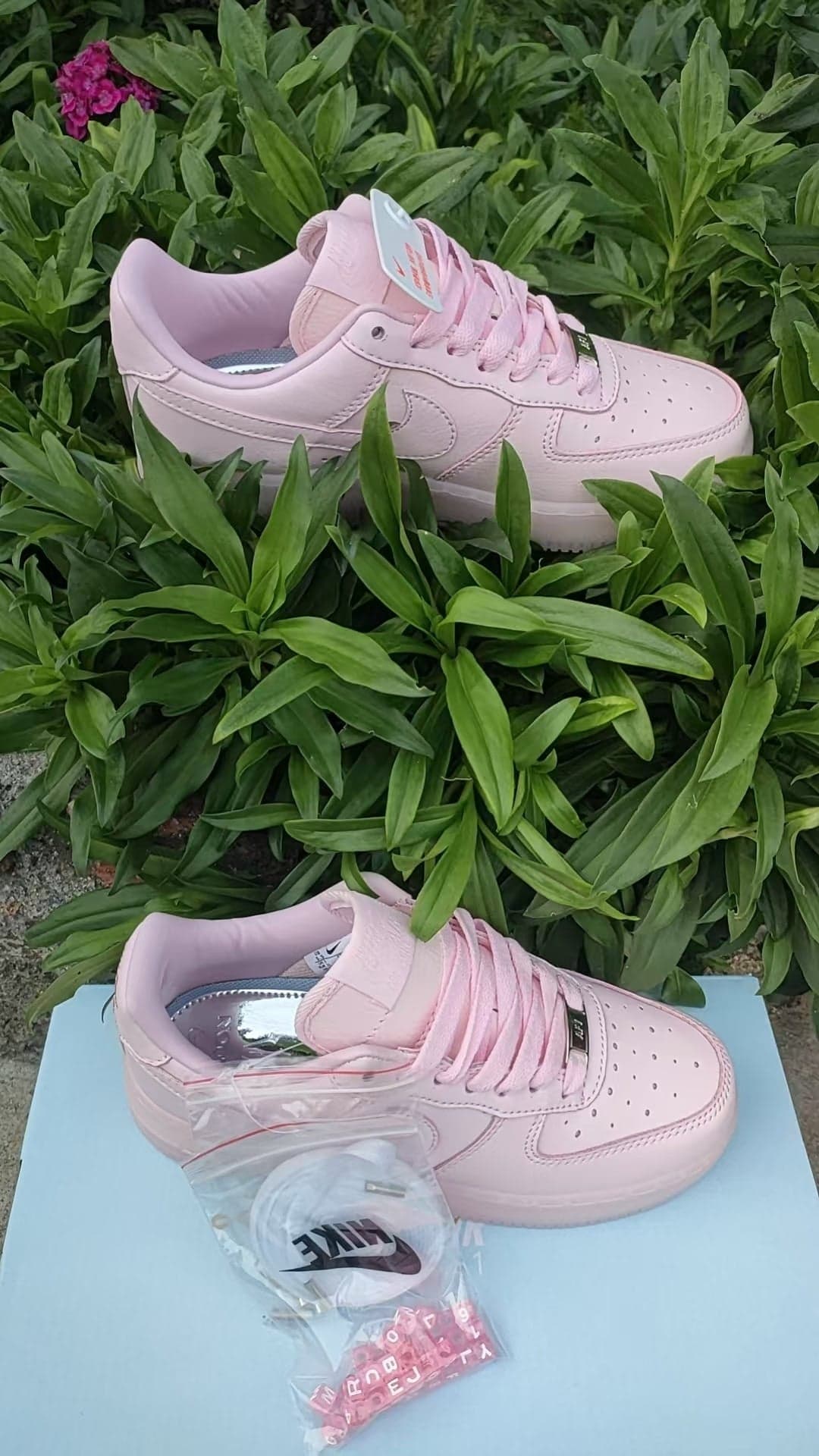 4fcf819a | UAE SPORT STORE Pastel pink Nike sneakers with laces and a tag, resting on a light blue box among green plants.