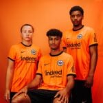 4n3X7Y7jq6z0VVL | UAE SPORT STORE Three athletes in bright orange soccer jerseys pose against an orange backdrop, two standing and one seated in the center.